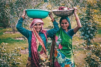 Two women wearing colorful traditional dresses smile outdoors, carrying baskets on their heads. HD wallpaper