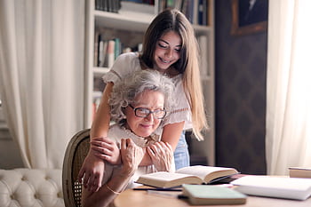 A heartwarming moment of a granddaughter embracing her grandmother as she reads a book together indoors. HD wallpaper