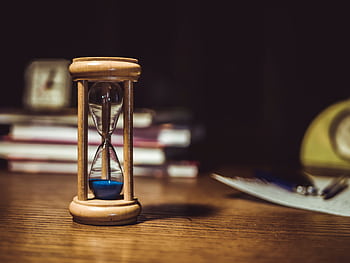 A close-up of a wooden hourglass with blue sand on a wooden desk, symbolizing time and patience. HD wallpaper