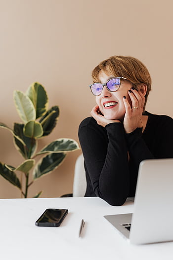 Cheerful woman at desk with laptop, smart, and plant, embodying a modern work lifestyle. HD phone wallpaper