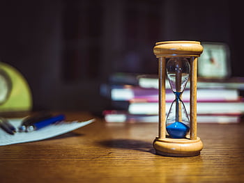 Close-up of a wooden hourglass on a desk, alongside books and pens, suggesting focused study or time management. HD wallpaper