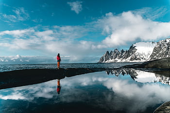 A person stands by a scenic snowy coastline reflected in still waters, under a vibrant blue sky. HD wallpaper