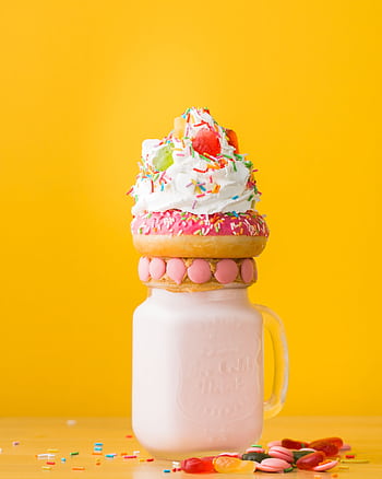 Vibrant milkshake adorned with a donut, whipped cream, and candy against a yellow background. HD phone wallpaper