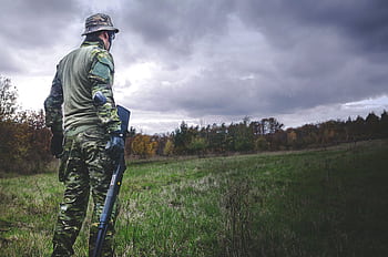 A soldier in camouflage with a rifle stands in a grassy field under a cloudy sky. HD wallpaper
