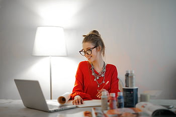 Smiling woman in red using laptop and art supplies at home office desk. HD wallpaper