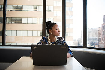 Confident woman working on a laptop in an office with large windows and city view. HD wallpaper