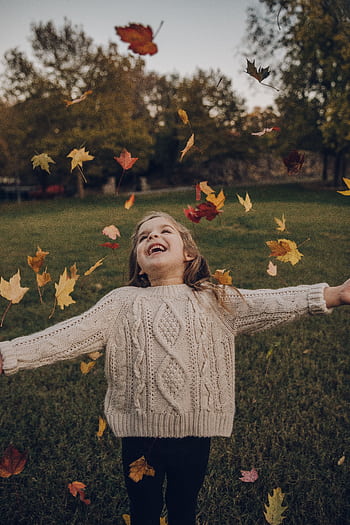 A young girl enjoys a playful moment surrounded by vibrant autumn leaves in a park. HD phone wallpaper