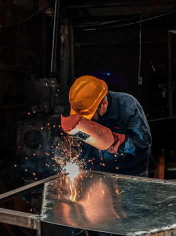 A welder wearing protective gear works on metal in an industrial workshop, surrounded by sparks. HD phone wallpaper
