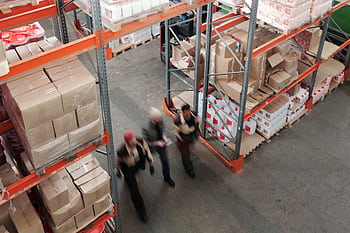A high angle view of workers in motion in a busy warehouse with shelves full of boxes and packages. HD wallpaper