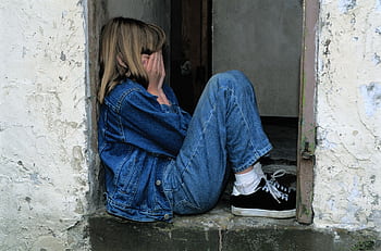 A young girl in denim sits alone in a doorway looking distressed, conveying loneliness. HD wallpaper