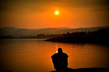 A person sits alone by a tranquil lake, with a stunning sunset in the background. HD wallpaper