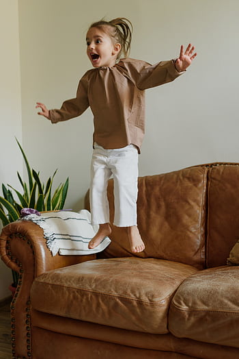 Happy child energetically jumping on a comfortable leather sofa indoors. HD phone wallpaper