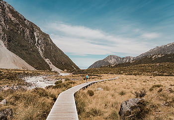 A hiker on a tranquil boardwalk path in New Zealand's mountainous landscape under a clear sky. HD wallpaper