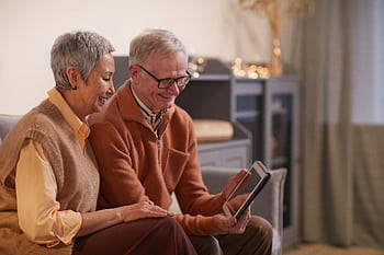 A cheerful senior couple enjoying a video call on a tablet in their cozy living room. HD wallpaper