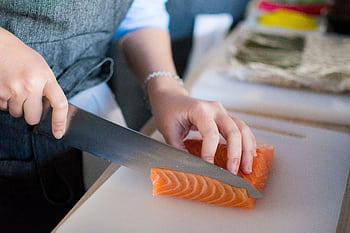 Close-up of a chef's hands slicing fresh salmon for sushi on a cutting board indoors. HD wallpaper