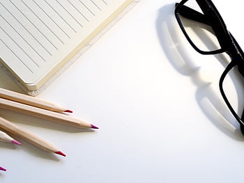 A clean desk setup showing a lined notebook, pencils, and black glasses on a white surface. HD wallpaper