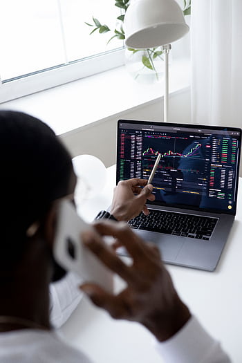 A businessman using a smart while analyzing stock market graphs on a laptop indoors. HD phone wallpaper