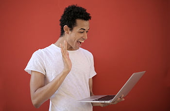 Young man making a cheerful video call on a laptop with a red background, expressing joy and surprise. HD wallpaper