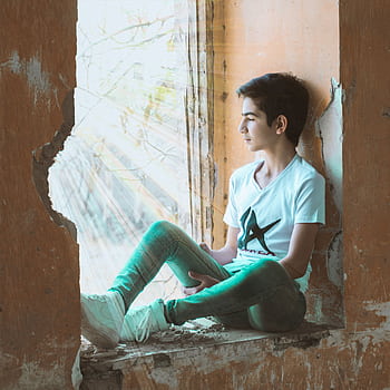 Young boy sitting in a sunlit window, wearing casual attire, gazing thoughtfully. HD phone wallpaper