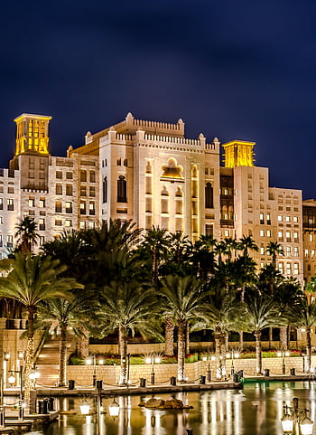 A stunning night view of a luxurious hotel with palm trees reflecting on water in Dubai. HD phone wallpaper
