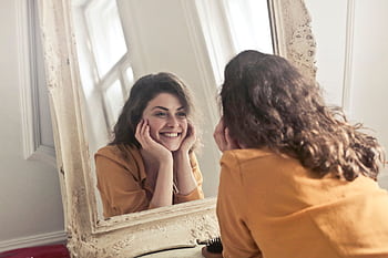 A cheerful woman smiles at her reflection in a vintage-style mirror, exuding positivity and warmth. HD wallpaper