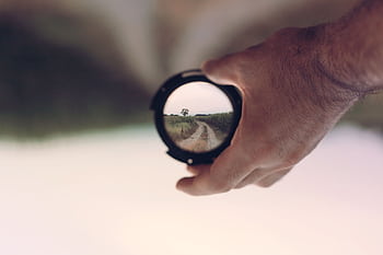A hand holds a camera lens revealing a clear path through nature, symbolizing vision and focus. HD wallpaper