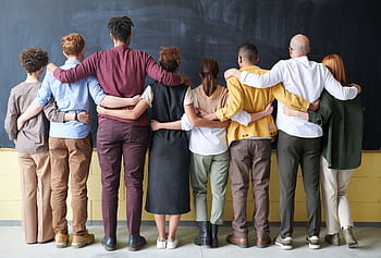 A diverse group of adults in casual outfits hugging in front of a chalkboard, symbolizing teamwork. HD wallpaper