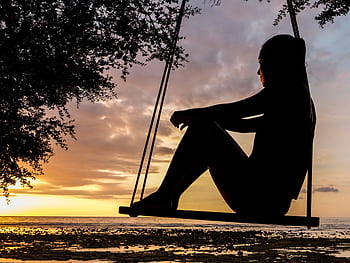 A serene sunset silhouette of a woman sitting on a swing by the seashore, evoking tranquility and reflection. HD wallpaper