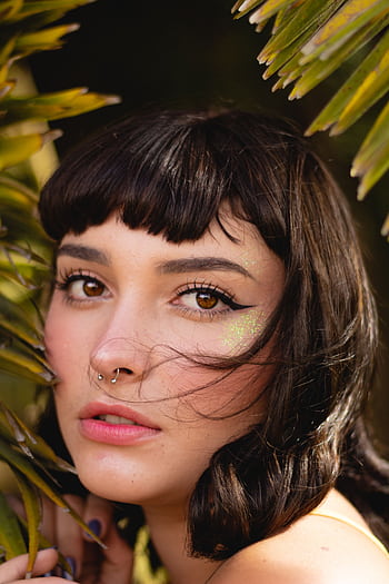 Stunning portrait of a young woman with brown eyes and septum piercing surrounded by tropical leaves. HD phone wallpaper