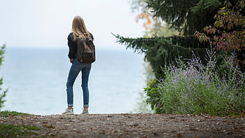 A young woman with a backpack stands on a scenic nature path overlooking a lake. HD wallpaper