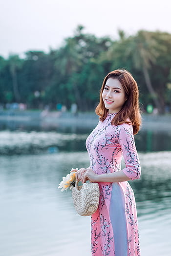 A young woman in a traditional pink dress smiles by a lake, holding flowers in a woven bag. HD phone wallpaper