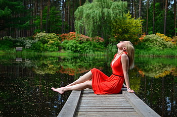A woman in a red dress enjoying a serene moment on a lakeside dock surrounded by lush greenery. HD wallpaper