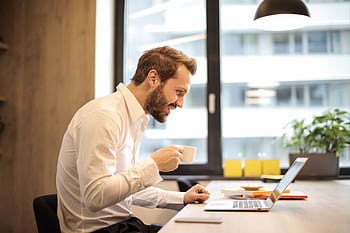 Man in office with coffee, smiling while working at laptop, captures the essence of remote work. HD wallpaper