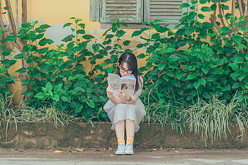 A young girl sits on a bench outdoors, immersed in a book with lush greenery around her. HD wallpaper