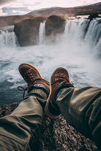 Person in hiking boots sitting at cliff's edge, overlooking breathtaking waterfall. HD phone wallpaper