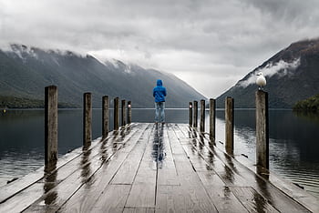 Person in blue jacket stands on a rainy dock overlooking serene lake and mountains. HD wallpaper
