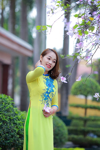 Young woman in ao dai smiles holding blossoms outdoors on a sunny day. HD phone wallpaper