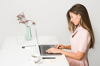 A woman in a pink dress working on a laptop at a modern, minimalist desk. HD wallpaper