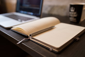 Open notebook with pen beside a laptop on a wooden desk, ideal for office or study themes. HD wallpaper