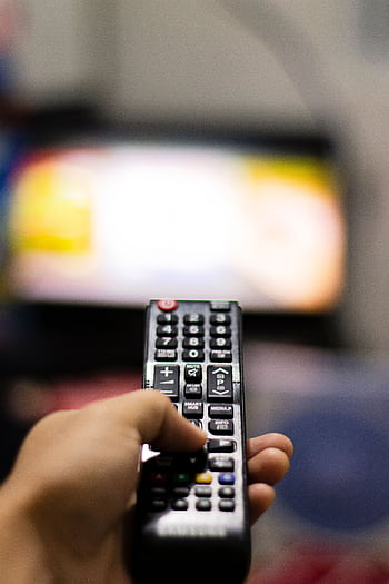 Close-up of a hand holding a remote control, watching TV indoors. Captures modern leisure and technology. HD phone wallpaper