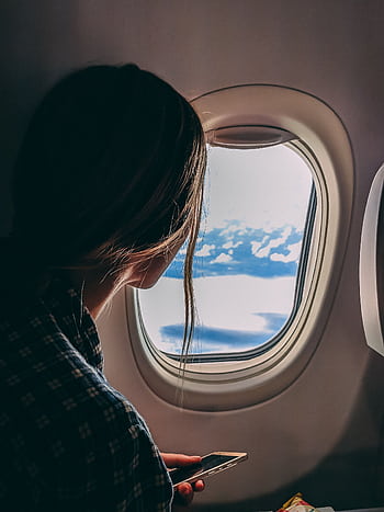 A woman looks out an airplane window, capturing a serene view of clouds and sky during a flight. HD phone wallpaper
