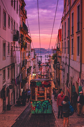Colorful tram navigating a narrow street in Lisbon during sunset with people and architecture. HD phone wallpaper