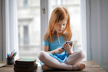 A young redhead girl in a blue dress using a smart indoors with books nearby. HD wallpaper