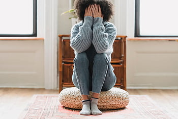 A woman sits on a pouf in a cozy home, face covered, conveying a moment of reflection and introspection. HD wallpaper
