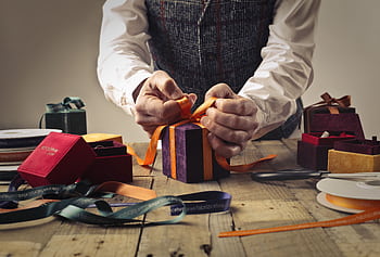 A person wraps a gift with orange ribbon on a rustic wooden table, surrounded by boxes. HD wallpaper