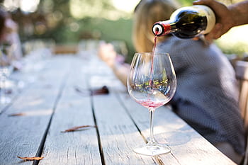 A hand pouring red wine into a glass on an outdoor table, creating a relaxed atmosphere. HD wallpaper