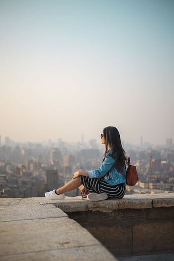 A young woman in casual attire sits on a rooftop terrace, viewing a sprawling cityscape in daylight. HD phone wallpaper