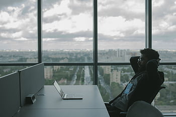 A man sits in an office with hands on head in front of a laptop, overlooking a cityscape. HD wallpaper