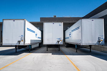 Three white cargo trailers parked at an industrial shipping dock under clear blue skies. HD wallpaper