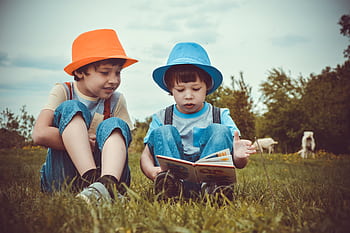 Two young boys enjoying a book together outdoors on a sunny day, wearing vibrant hats. HD wallpaper
